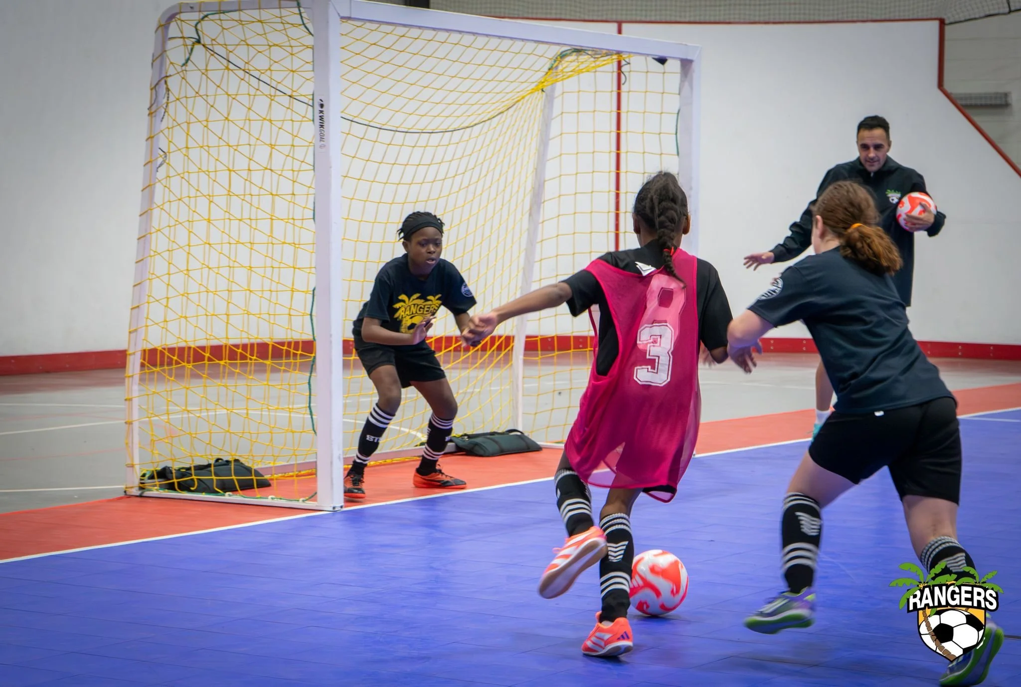 Kids playing indoor soccer, with one girl in pink kicking a ball towards the goal, while a girl in black tries to block, and a man with a ball nearby in the background.