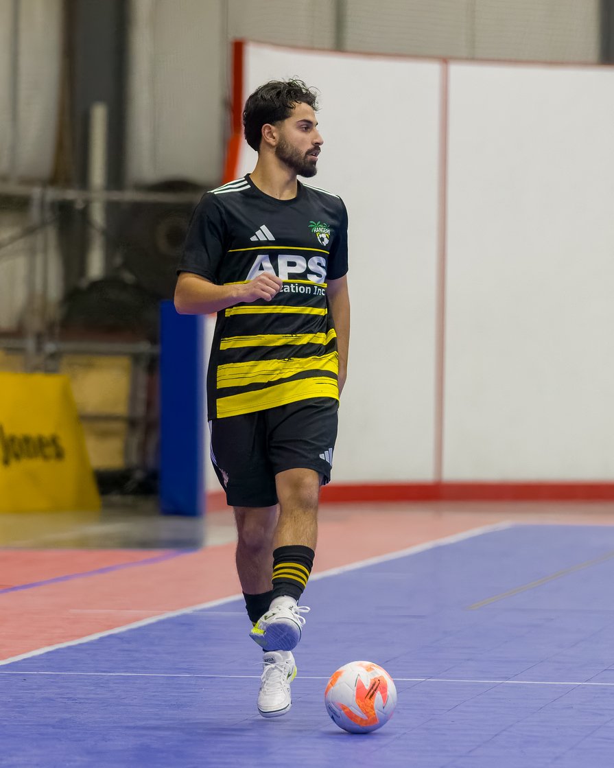 A man in a black and yellow soccer jersey and black shorts playing indoor soccer, standing on a blue court with a soccer ball at his feet.