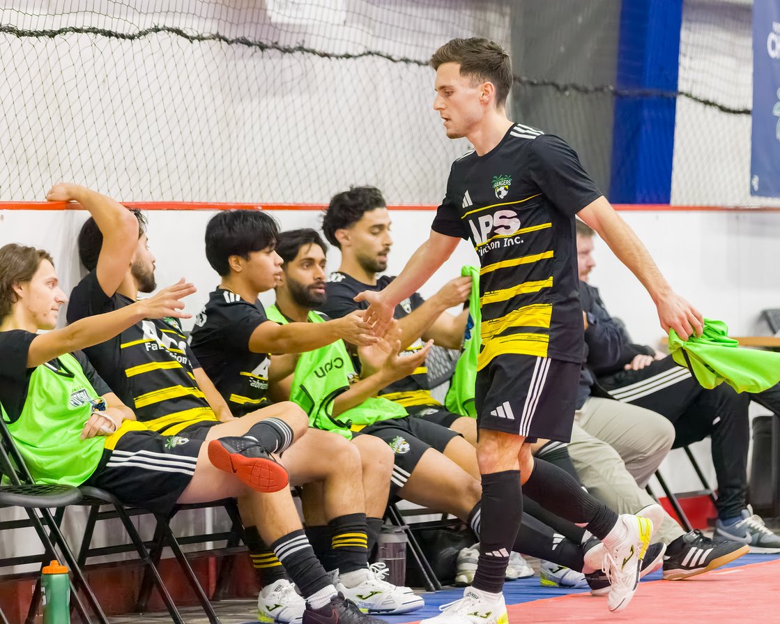 A soccer player is giving a high-five to a teammate sitting on the bench during a game, with other teammates sitting or standing nearby in an indoor sports facility.
