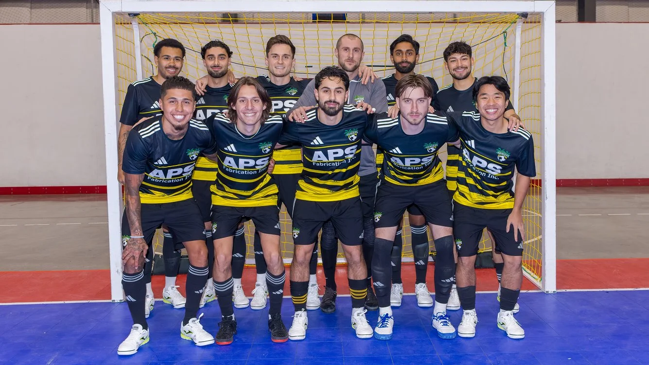 A group of nine men in dark blue and yellow futsal uniforms posing inside an indoor sports hall in front of a goal.