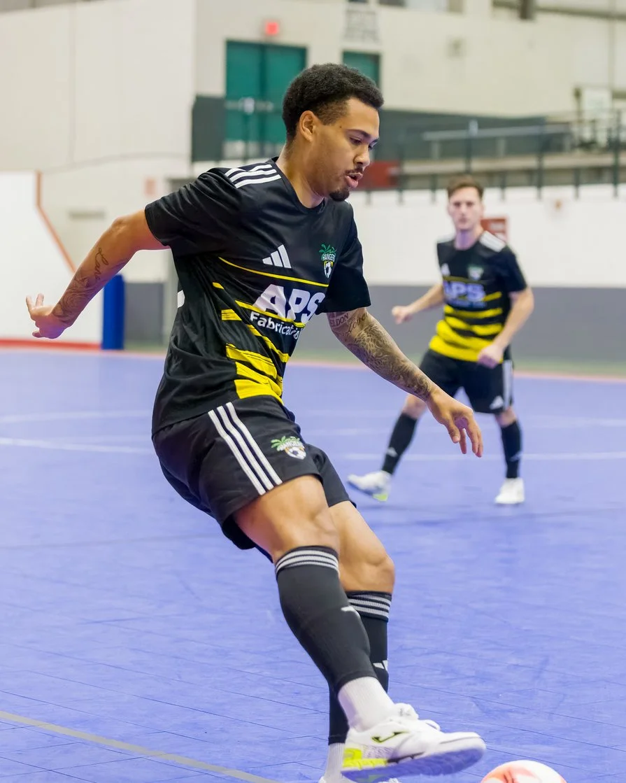 Two male soccer players in black and yellow uniforms playing indoor soccer on a blue court, with one player in the foreground and the other in the background.