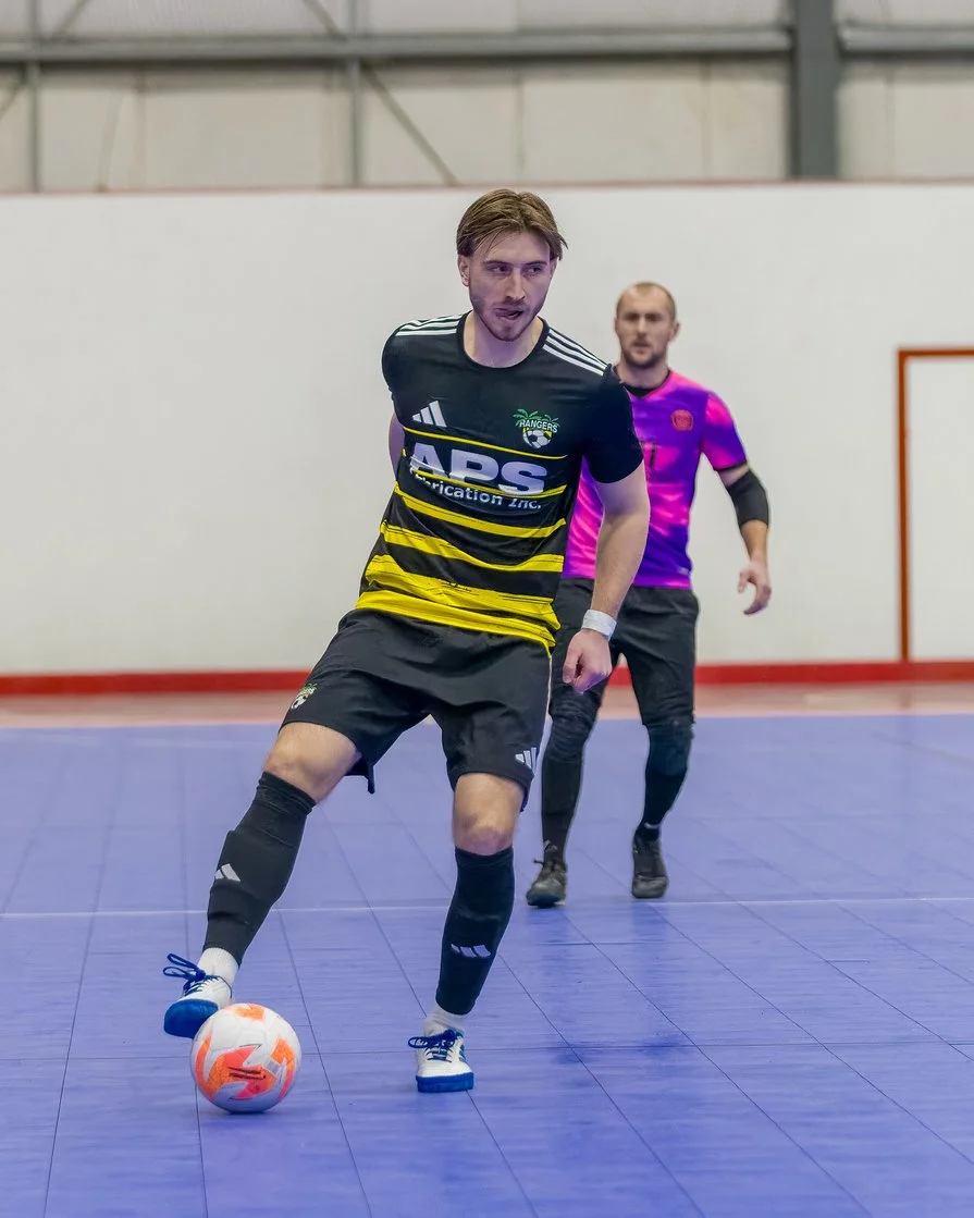A man in a black and yellow sports jersey and black shorts playing indoor soccer, dribbling a ball on a blue court with another man in a purple and black jersey running behind him.