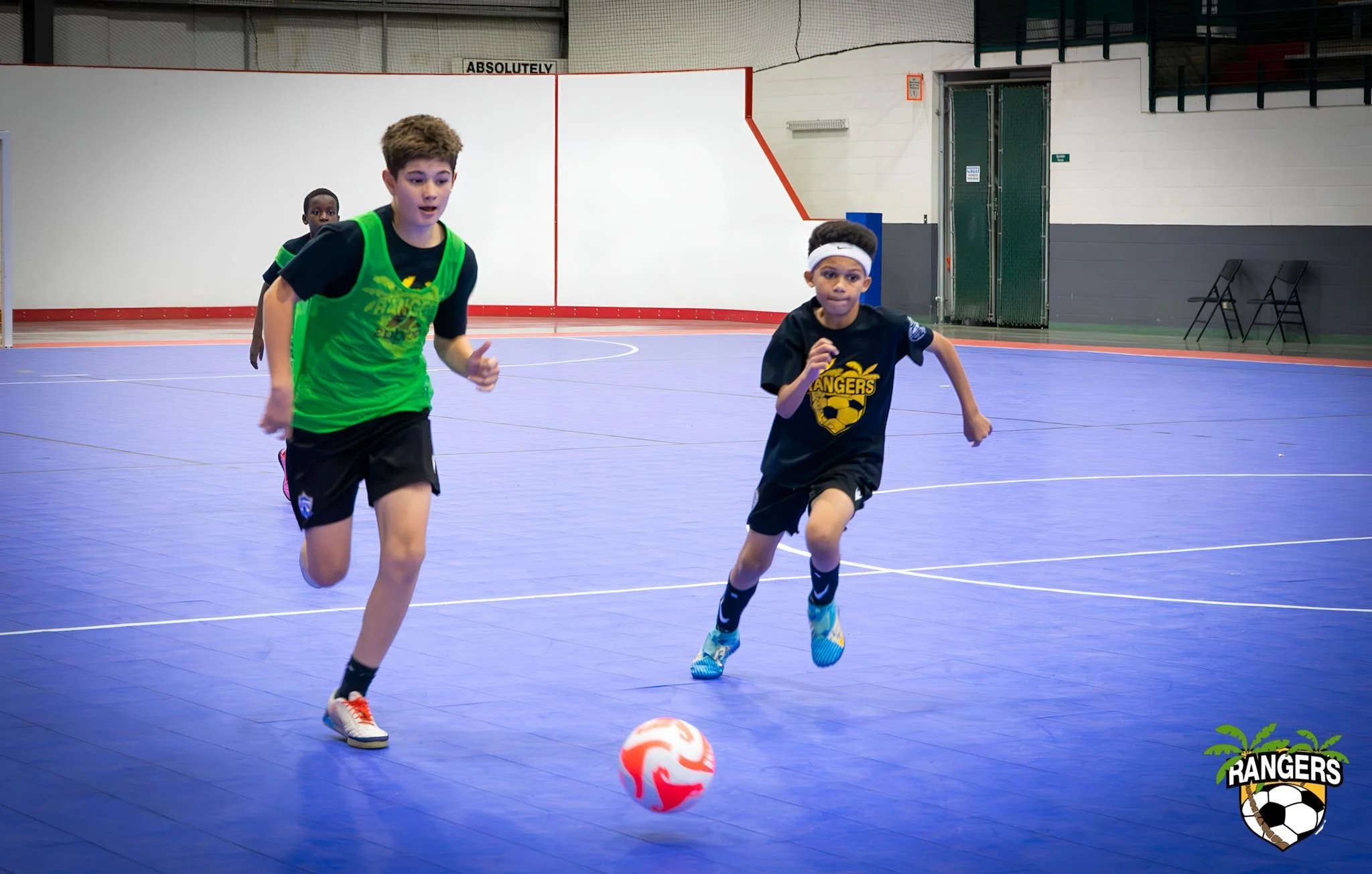 Two young boys are running and playing soccer on an indoor court. One boy is wearing a green and black jersey, and the other is wearing a black and yellow jersey with a white headband. There is a soccer ball in front of them, and the court has a blue