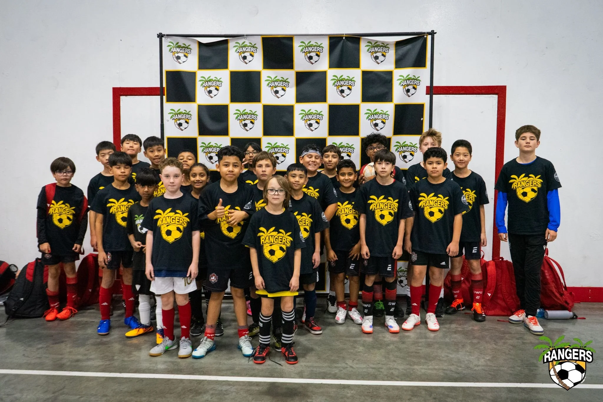 Group of young children in black t-shirts with a yellow soccer team logo posing for a team photo indoors, with some kids holding a soccer ball, in front of a black and white checkered backdrop with a sports team logo.