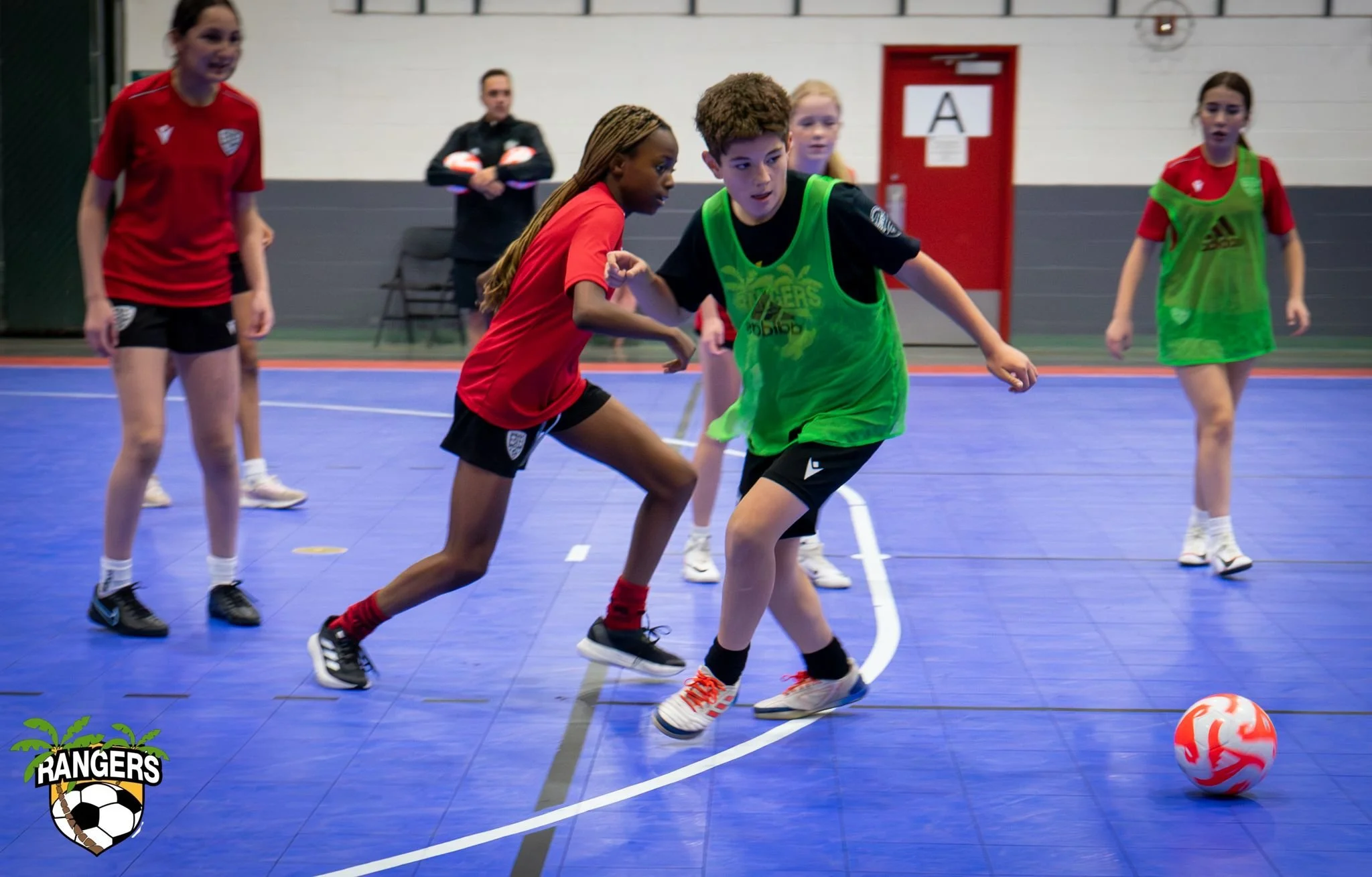 Children playing indoor soccer on a blue court, with one child in a green vest and others in red, chasing an orange and white soccer ball.
