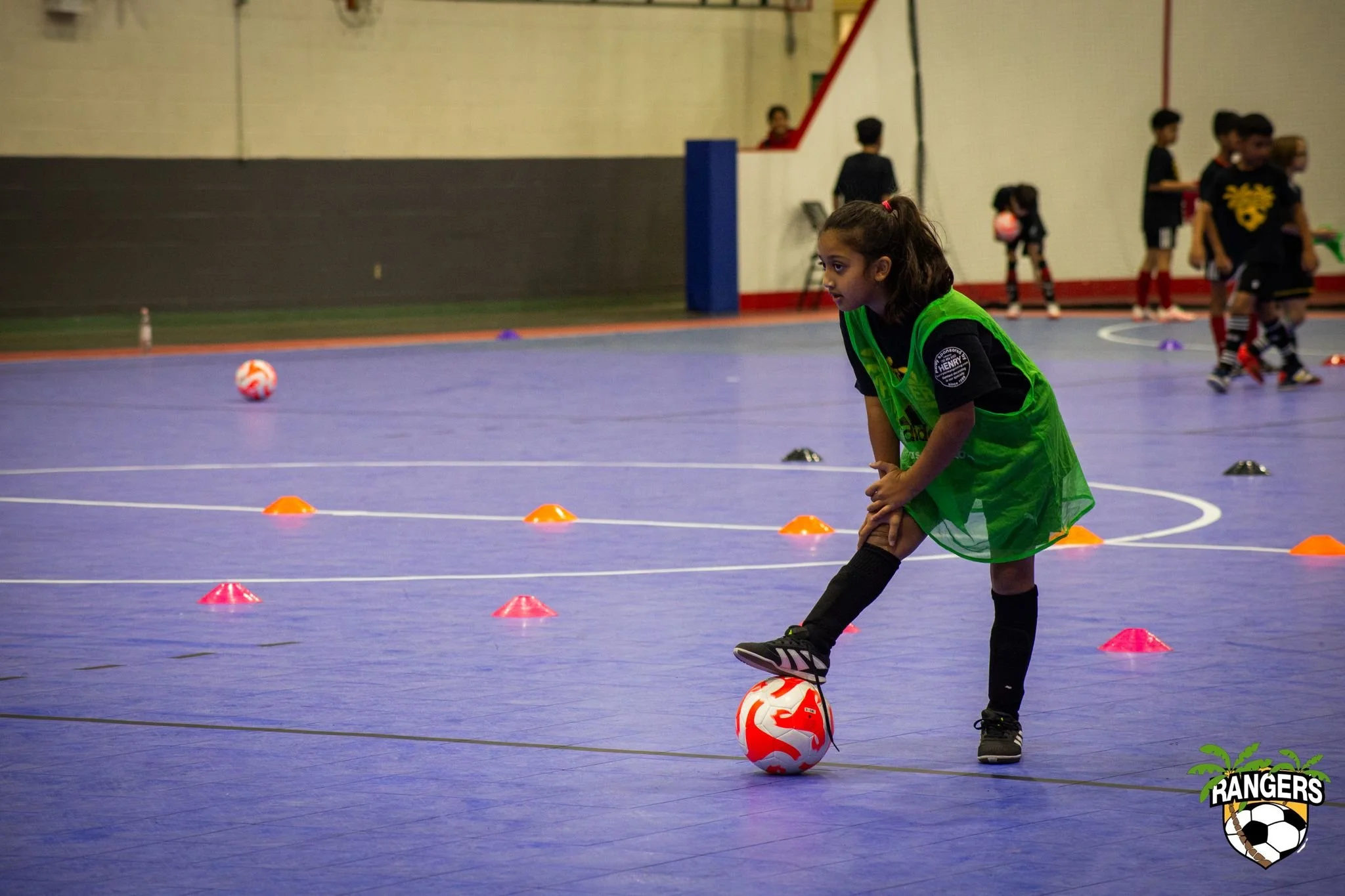 Young girl in a green sports bib practicing soccer drills on an indoor court, with several other children in the background, and the team logo 'RANGERS' with a soccer ball and palm trees in the bottom right corner.
