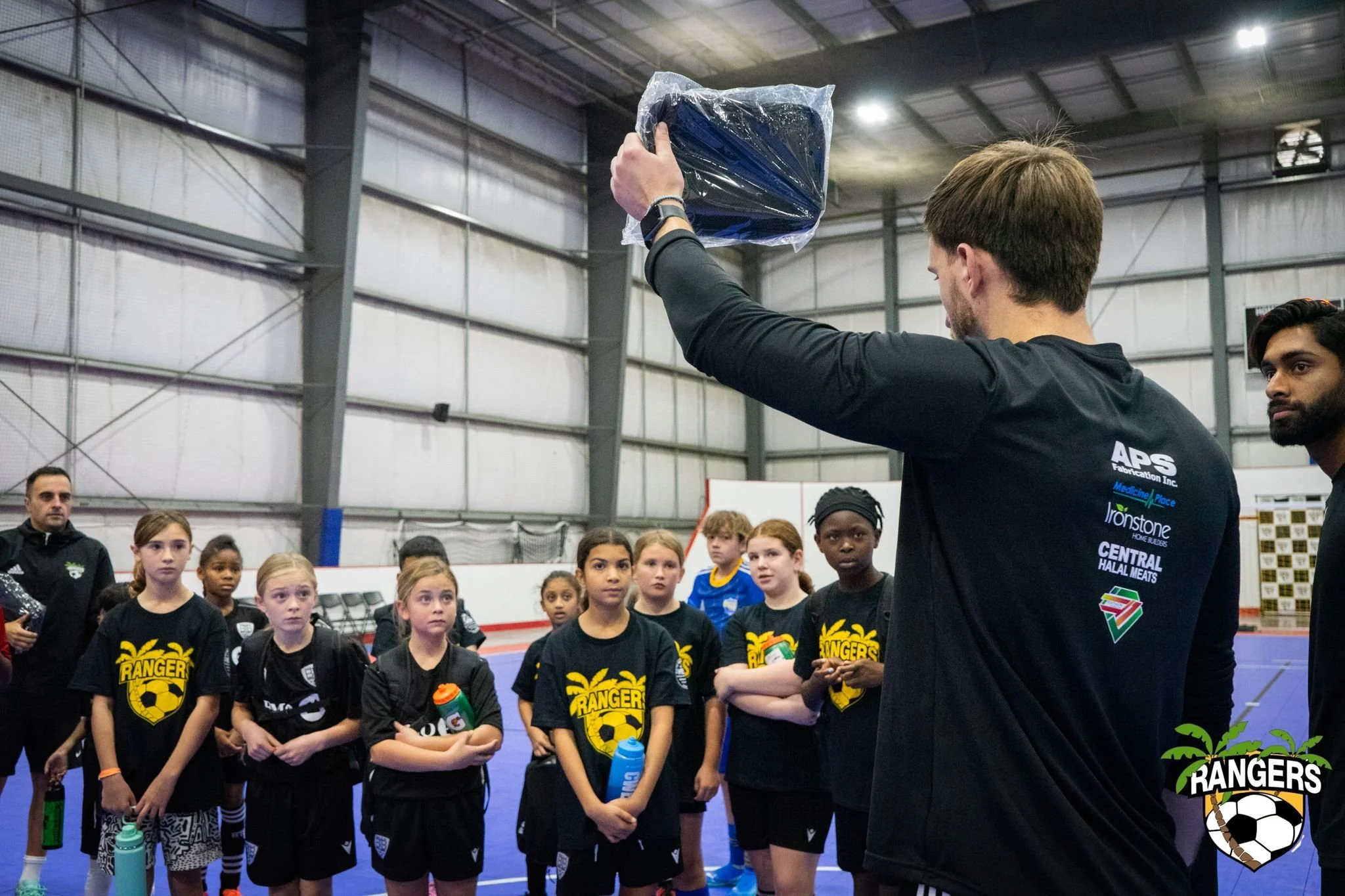 A coach is holding a wrapped item in front of a group of young female soccer players inside an indoor sports facility. The players are wearing black jerseys with a yellow logo, and some are holding water bottles. The facility has a curved metal roof 