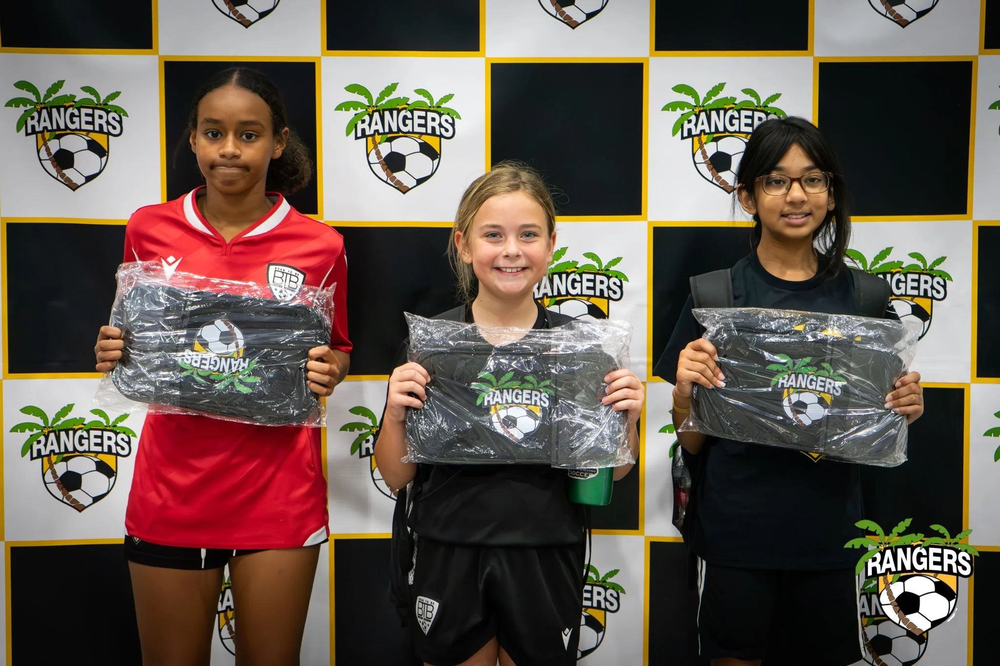 Three young girls standing in front of a Rangers soccer team backdrop, each holding clear plastic bags with the Rangers logo. The girl on the left wears a red soccer uniform, the girl in the middle wears a black sports outfit, and the girl on the rig