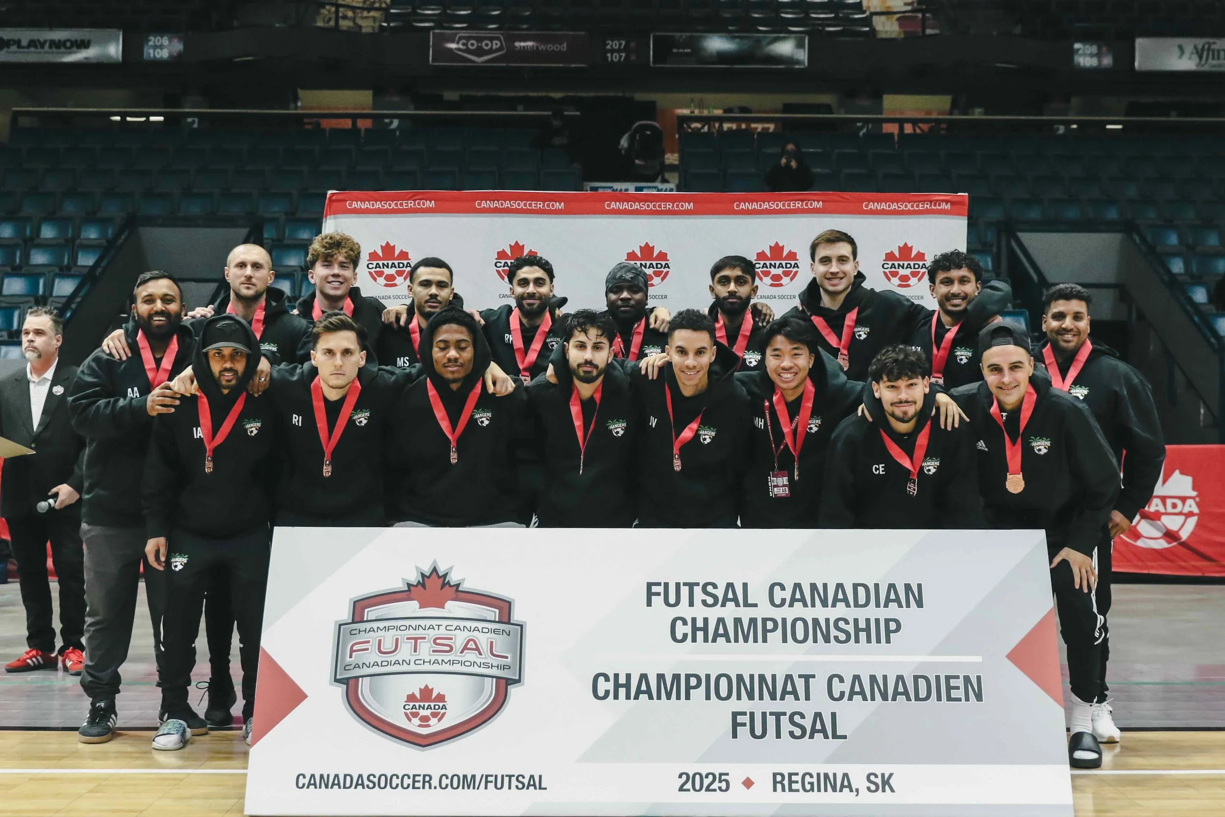 A group of smiling futsal players wearing black jackets with medals around their necks, standing behind a large banner that reads "Futsal Canadian Championship, Canadian Futsal, 2025, Regina, SK." They are in an indoor sports arena with some spectato