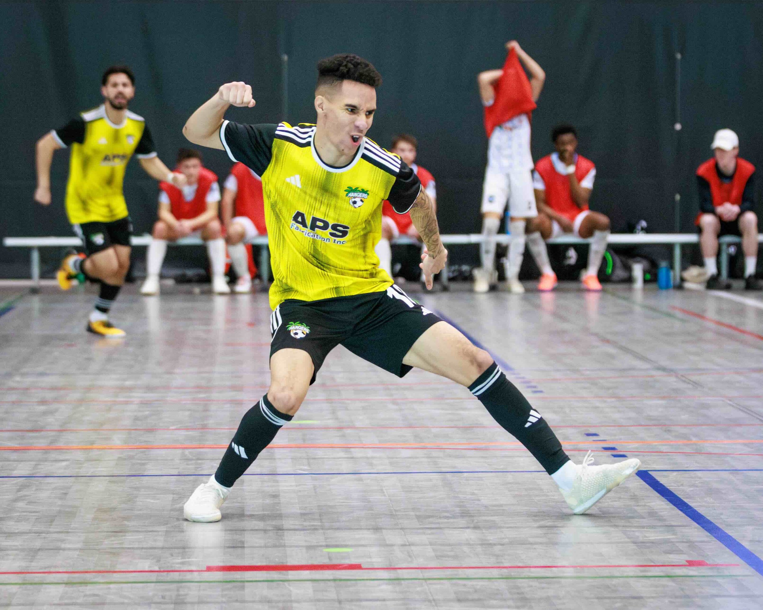 An indoor soccer player in a yellow and black uniform celebrates on a gym floor, with teammates and coaches sitting and standing in the background.