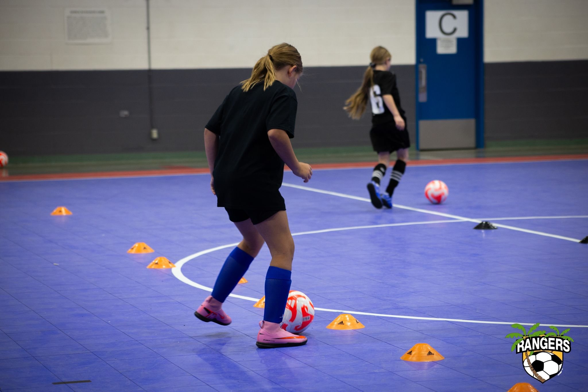 Two young girls practicing soccer in an indoor gymnasium, dribbling with soccer balls around orange cones, with a blue door and a logo in the bottom right corner that says 'Rangers' with a palm tree and soccer ball.