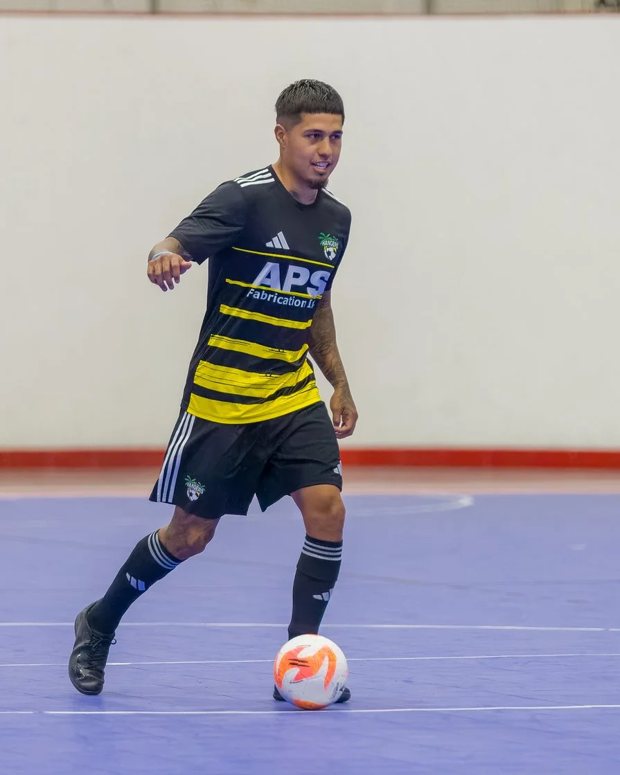 A young man in a black and yellow sports jersey and black shorts playing indoor soccer, controlling a white and orange soccer ball on a blue indoor court.