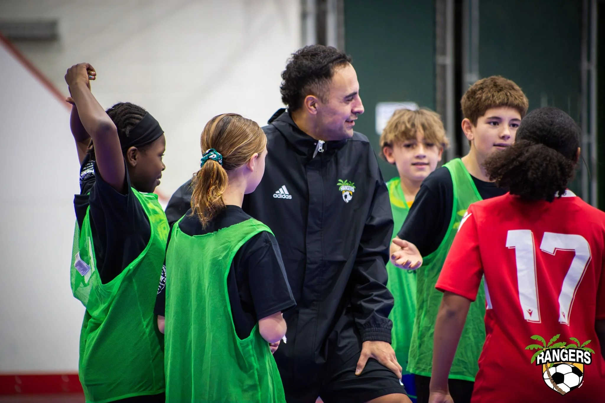 A coach or trainer talking to young soccer players during a practice session, with a logo that reads 'Rangers' on their jerseys.