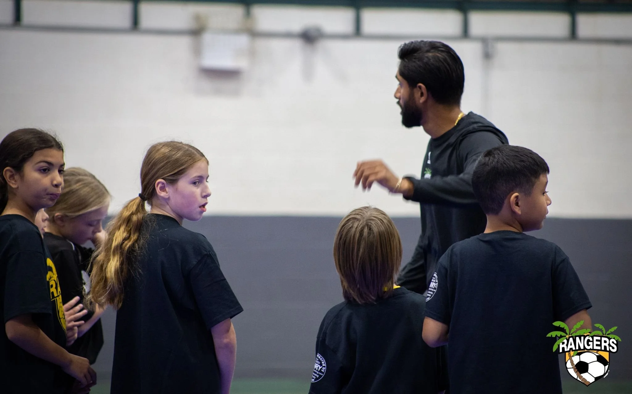 A coach speaking to a group of young children during a soccer practice indoors, with a logo of a soccer ball and the word 'RANGERS' in the bottom right corner.