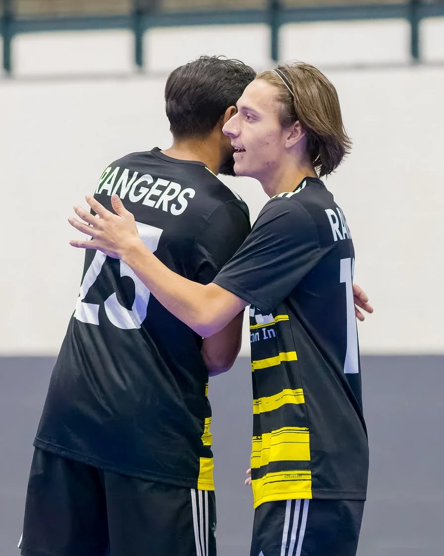 Two male athletes in black jerseys hugging inside an indoor sports arena.