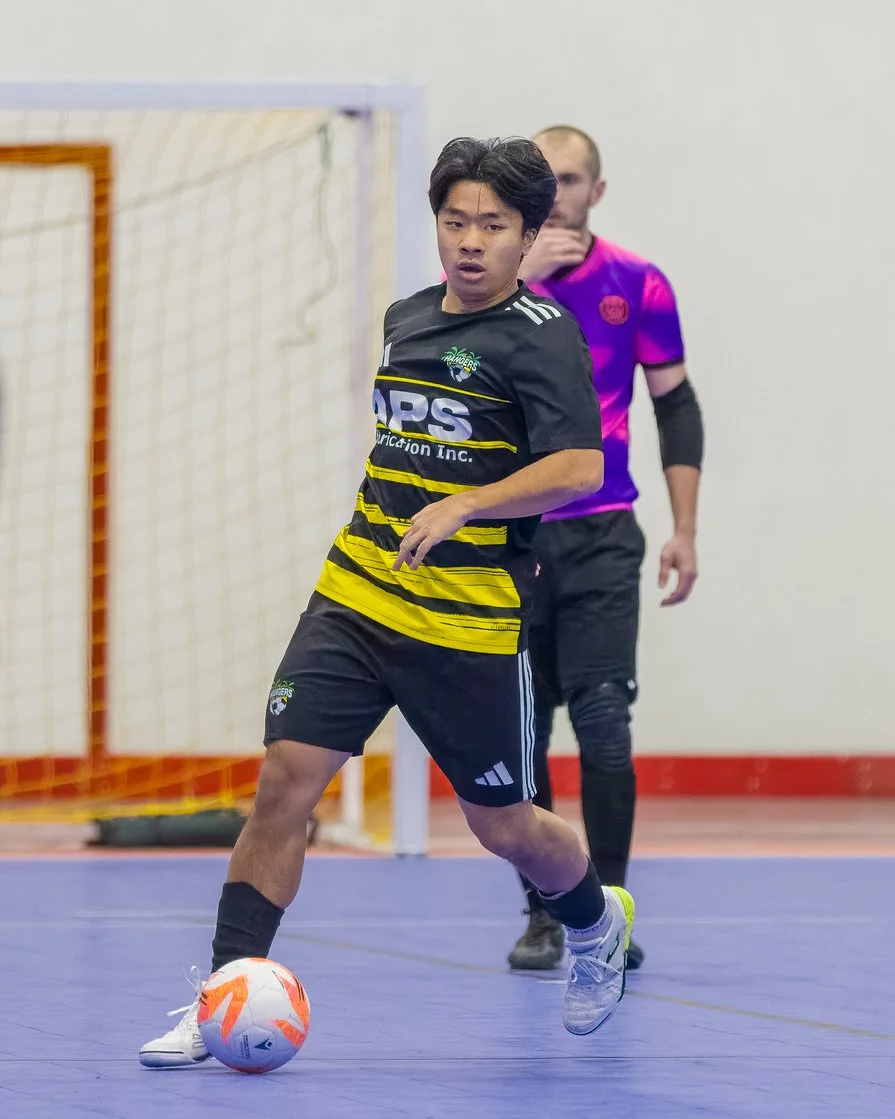 A young man playing indoor soccer, wearing a black and yellow jersey, black shorts, and white cleats, is about to kick a orange and white soccer ball. A referee or coach in a purple jersey stands in the background.