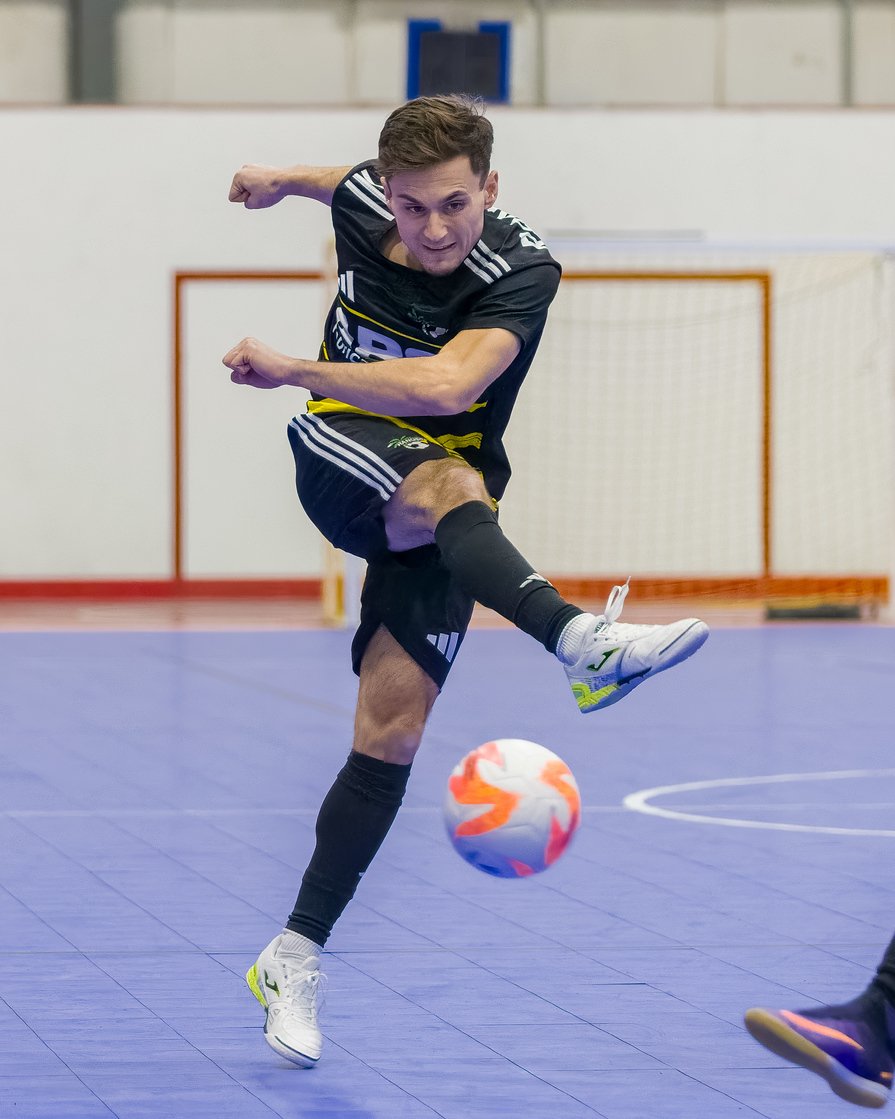 A young man with short brown hair wearing a black sports uniform with white and yellow accents is kicking a multicolored soccer ball inside an indoor sports facility.