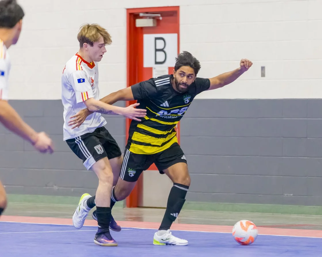 Two soccer players competing for ball in an indoor game, one in black and yellow jersey, the other in white jersey, with a red door and grey wall in the background.