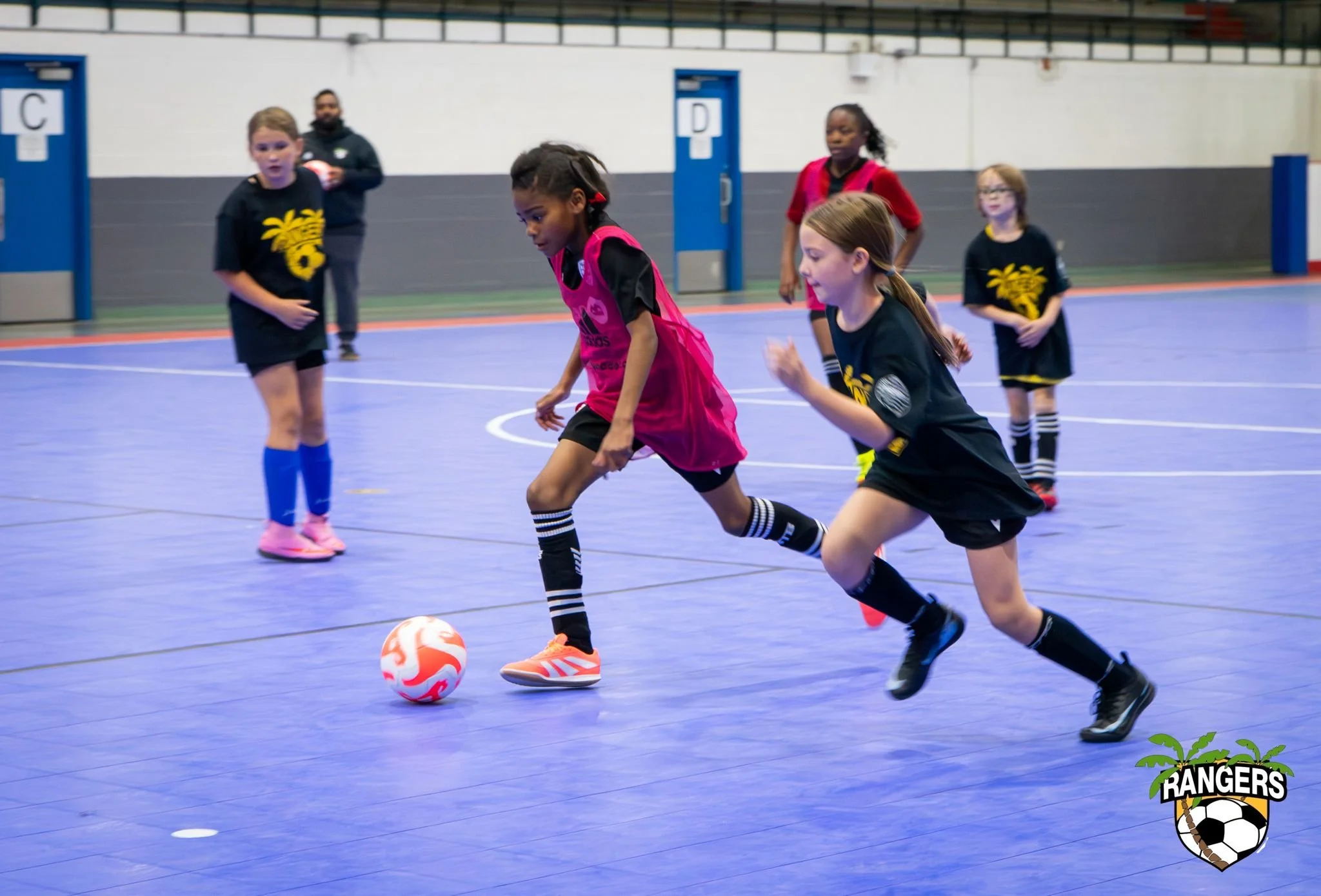 Young girls playing indoor soccer on a blue court, with one girl in a pink bib dribbling the ball while others watch or run nearby. The logo of Rangers soccer team is visible in the bottom right corner.