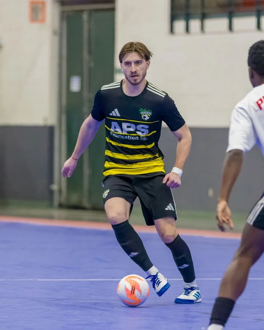 A man playing indoor soccer dribbling a ball, wearing a black and yellow uniform with Adidas shoes.