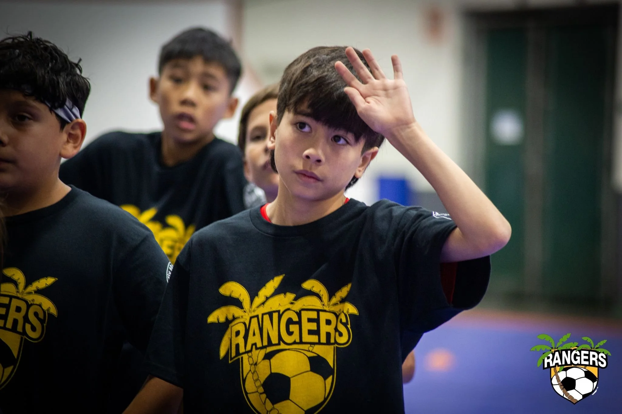 Young boy with dark hair in a black shirt with 'RANGERS' logo, raising his hand to his forehead during a soccer event with children in the background.