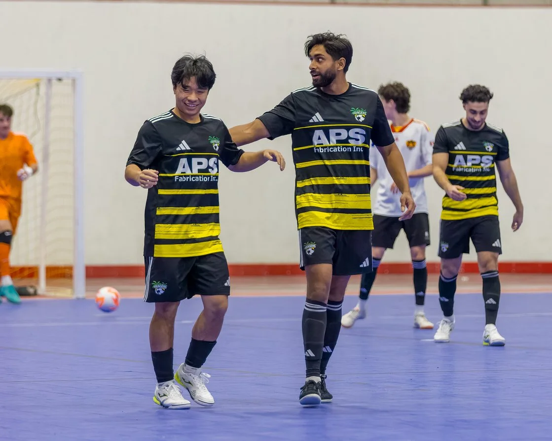 A group of soccer players in black and yellow jerseys celebrating on an indoor court with a purple floor, with one player smiling and others walking behind him.