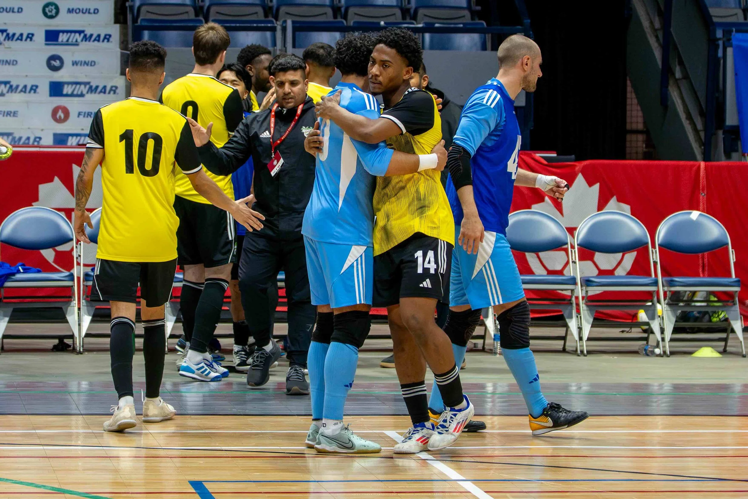 Two futsal players hugging on an indoor court after a match, with other players and staff in the background.