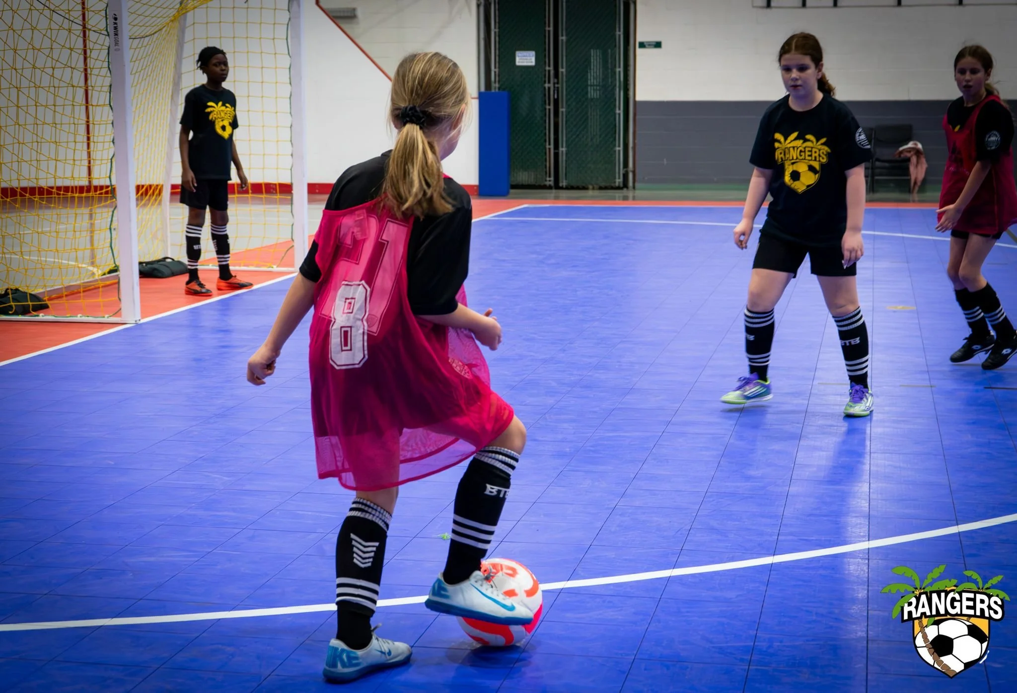 Young girls playing indoor soccer on a blue court, with a girl in a pink jersey and black shorts in the foreground, standing with a soccer ball at her feet. Other players and a goal are visible in the background.