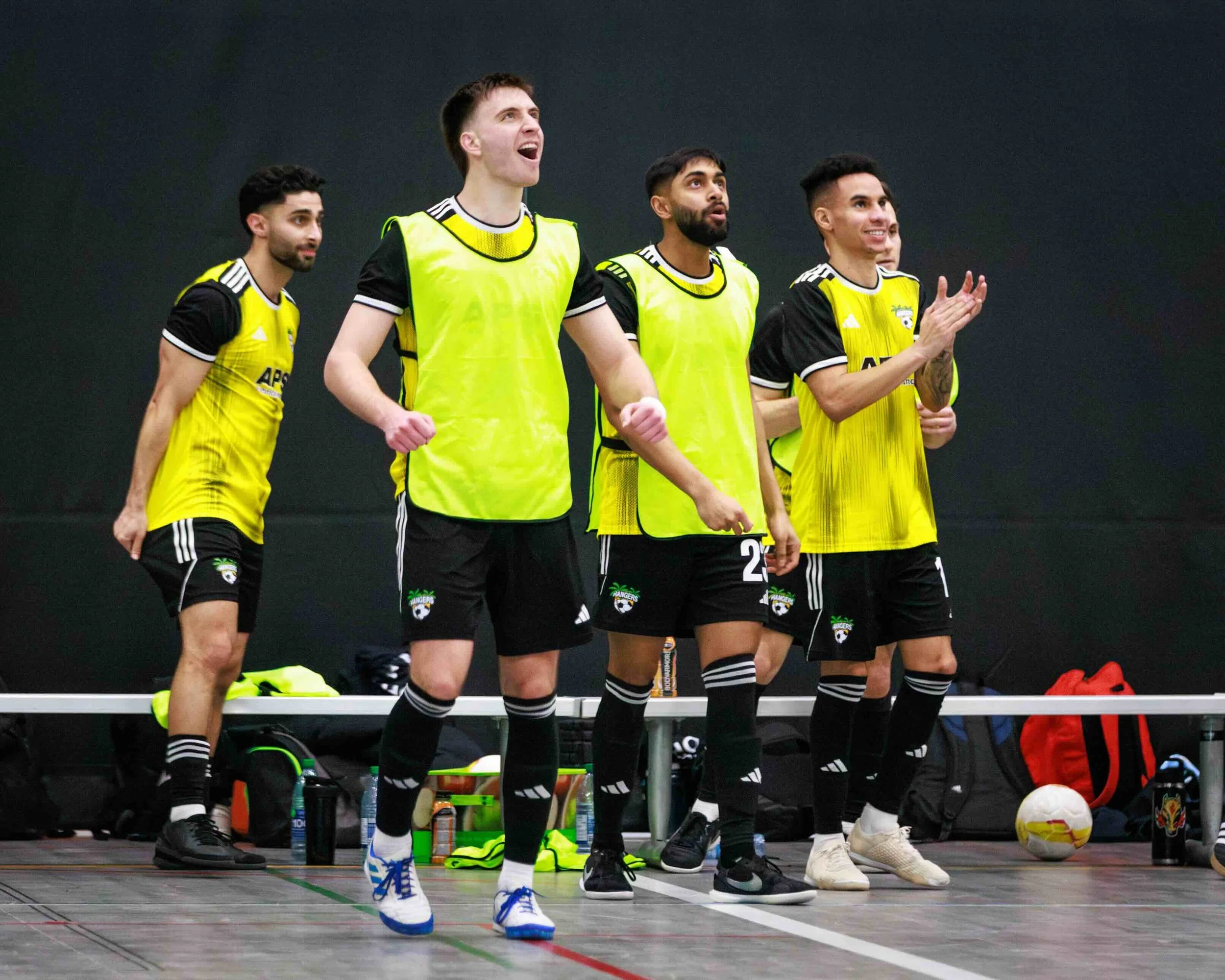 Group of male indoor soccer players wearing yellow bibs, standing on the sideline, with some clapping and cheering, with soccer equipment and bags in the background.