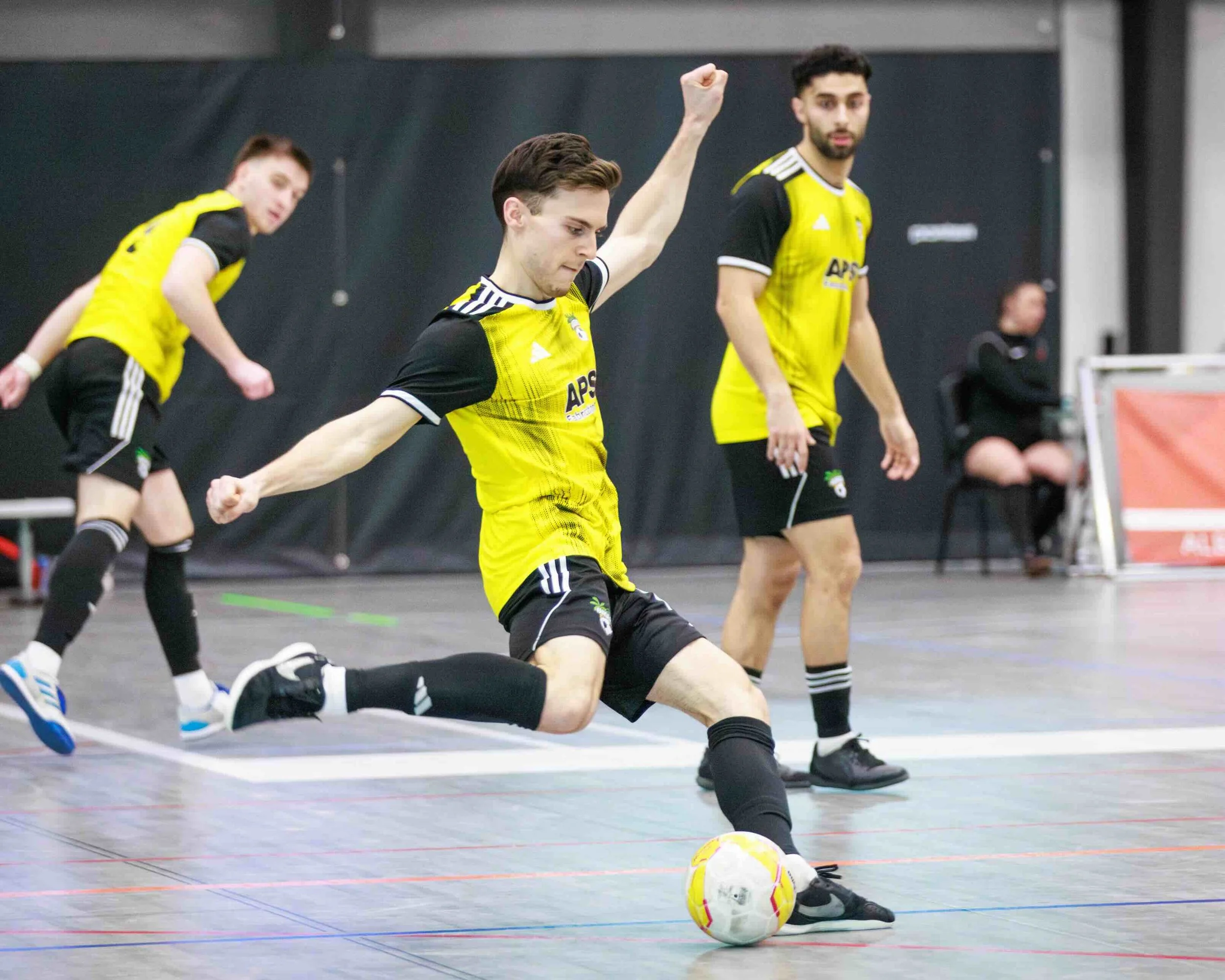 Young man in yellow and black soccer uniform kicking a yellow and white indoor soccer ball on a court, with two other players in similar uniforms and a woman sitting in the background.