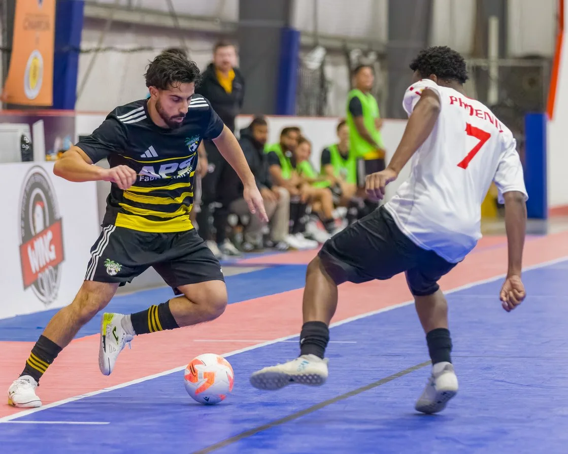 Two men playing indoor soccer, one in a black and yellow uniform dribbling the ball, and the other in a white uniform with red number seven defending, with players and coaches seated in the background.