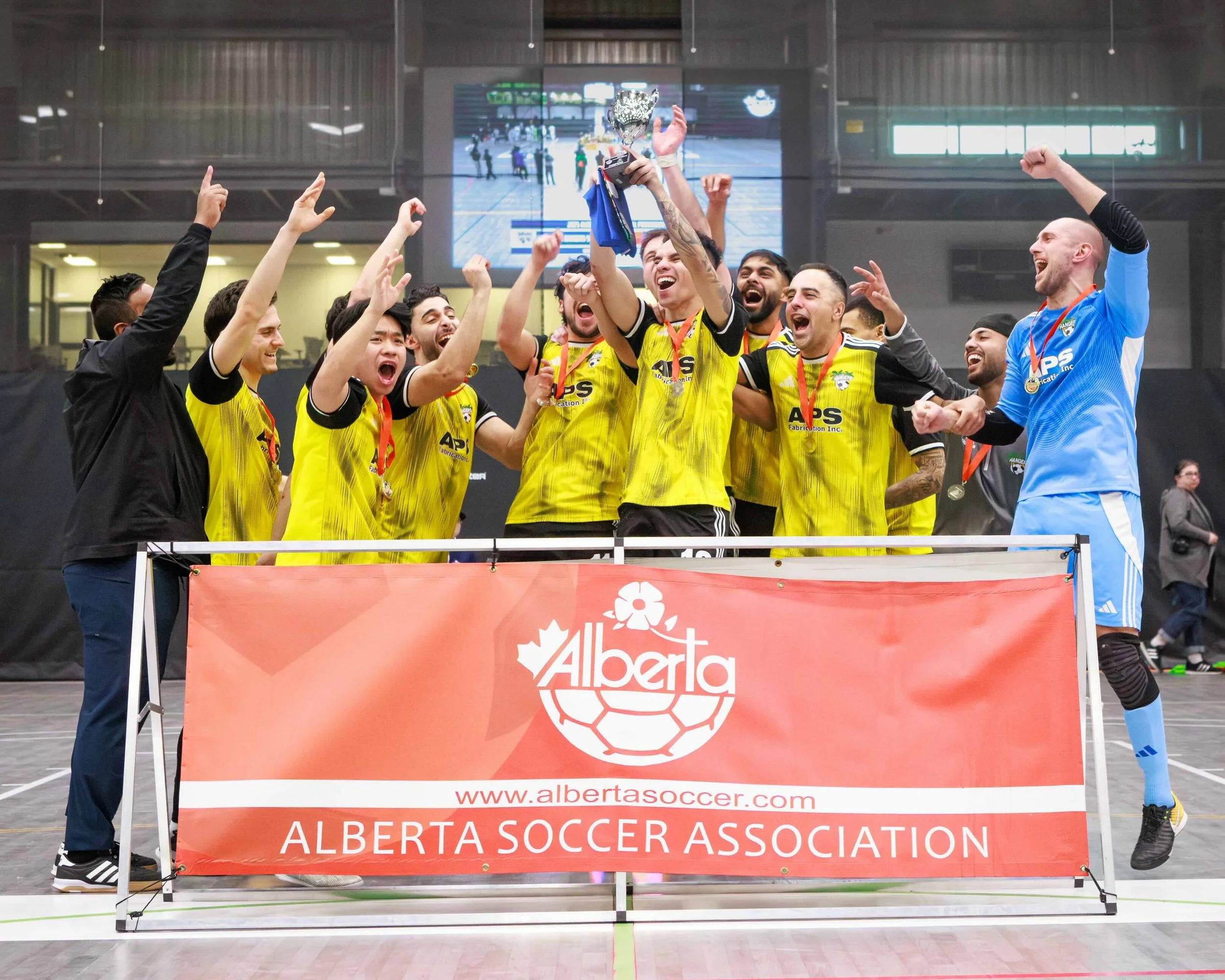 A men's soccer team in yellow jerseys celebrating victory and lifting a trophy on an indoor soccer court. The team is behind a red banner that reads 'Alberta Soccer Association' with a website link. The players are cheering and have medals around the