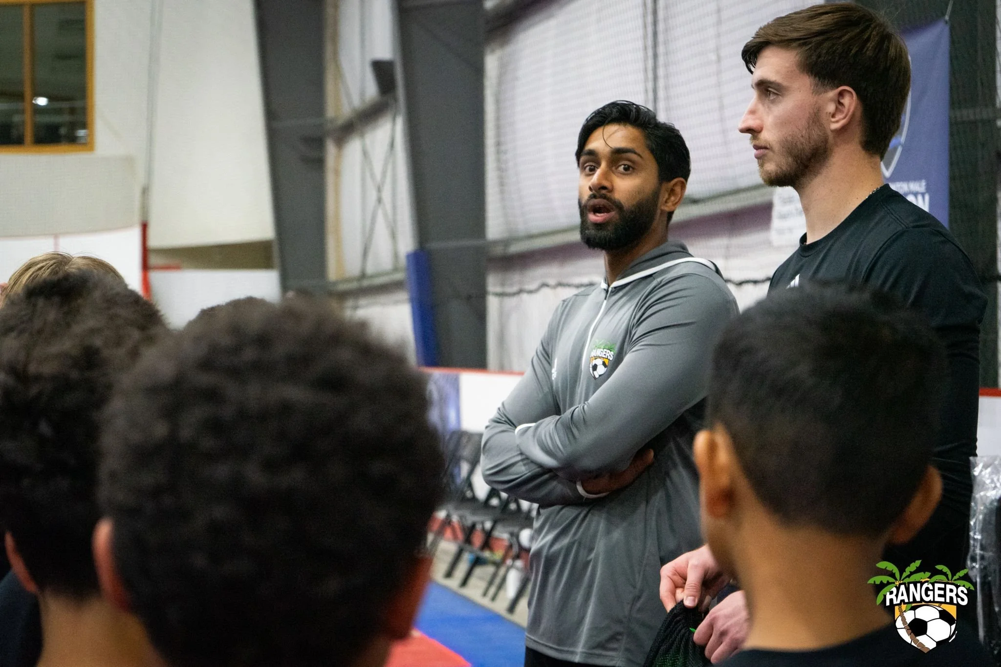 Two men standing and speaking to a group of young people in a sports facility. One of the men is wearing a gray jacket with a 'Rangers' logo. The audience is mostly seen from behind.