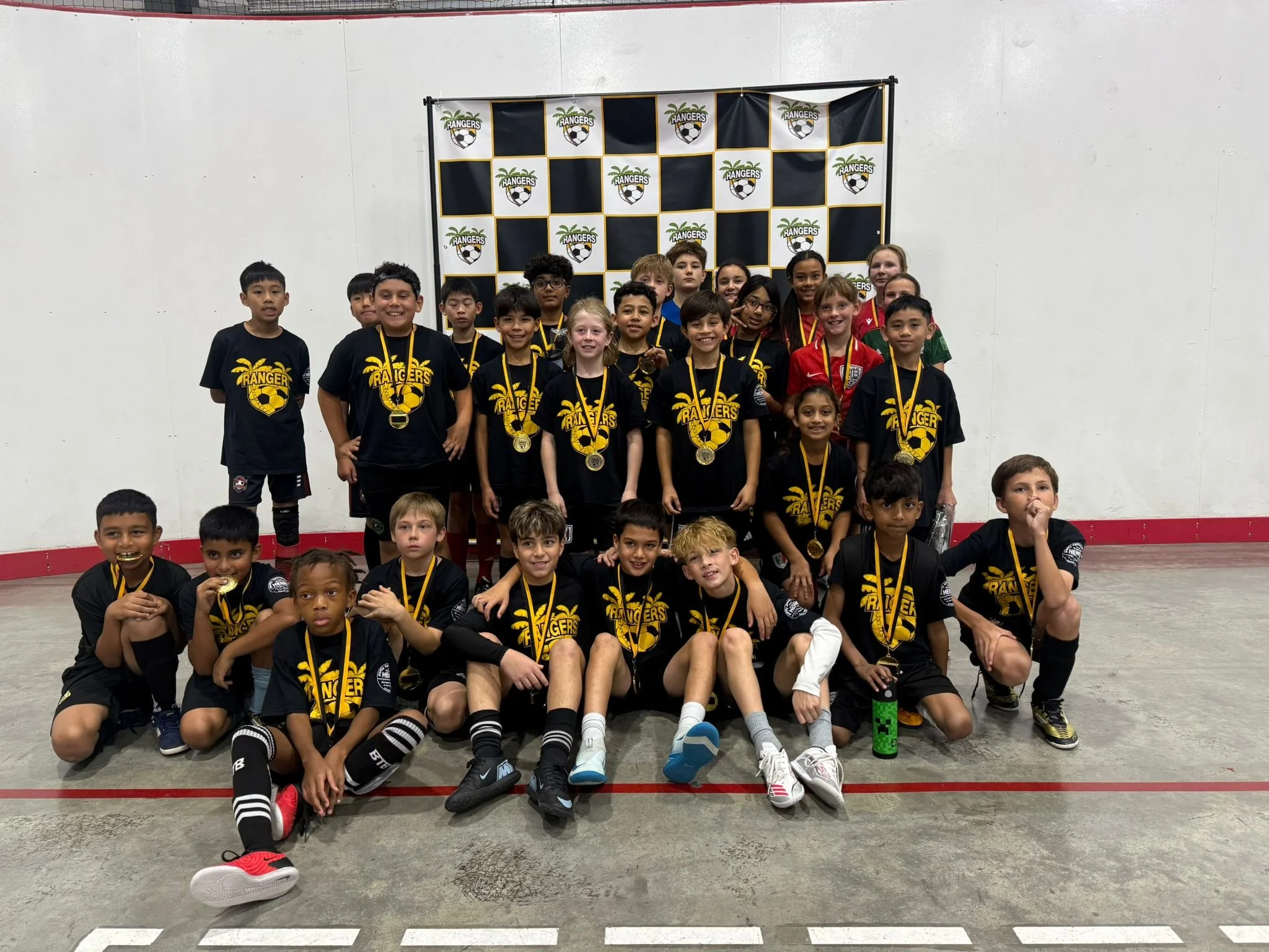 Children in black soccer jerseys with gold medals posing for a group photo indoors, with a black and white checkered backdrop featuring a soccer ball and the word 'Rangers' behind them.