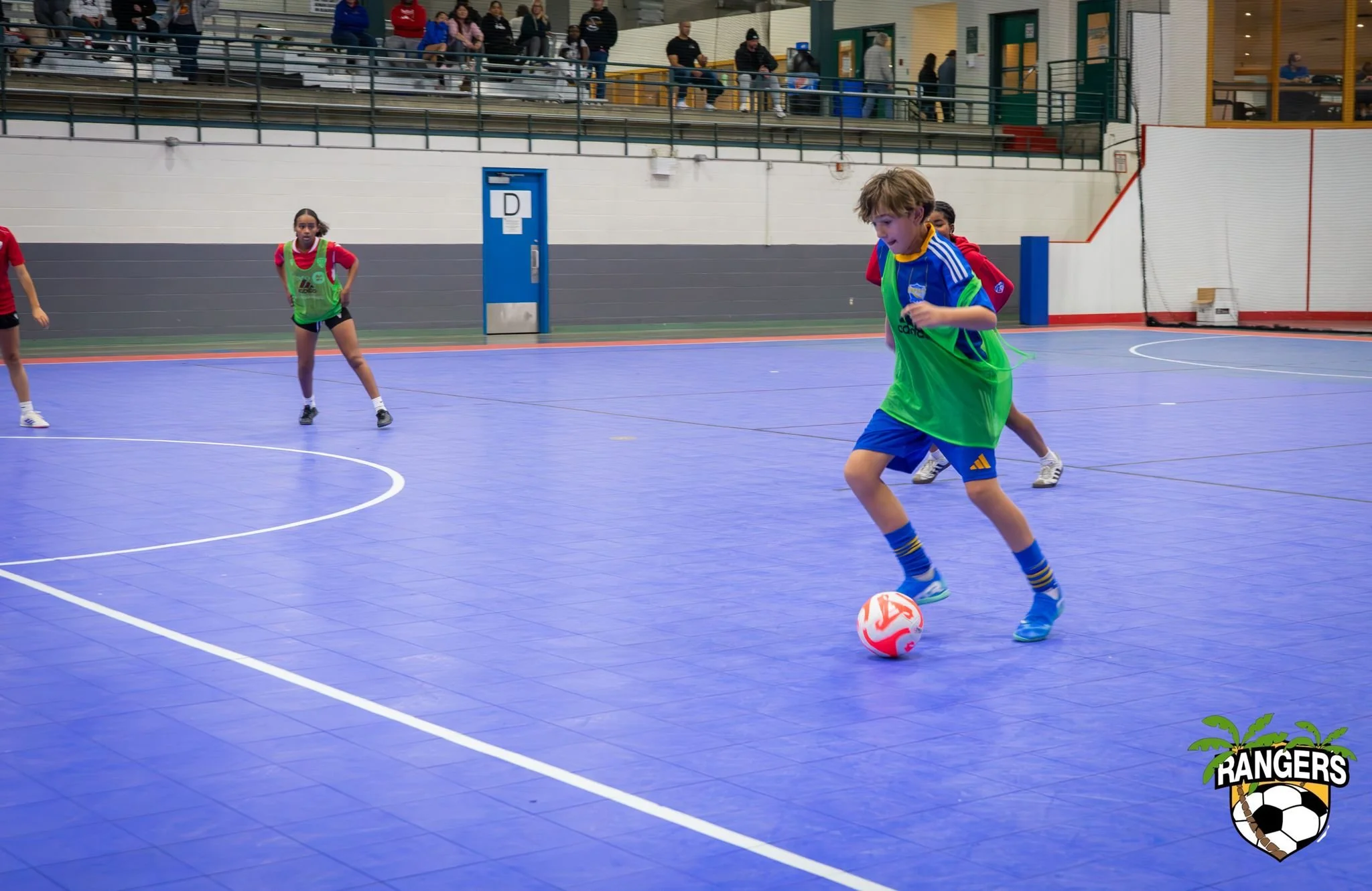 Children playing indoor soccer on a blue court, with some children in green jerseys and others in red and blue, and spectators watching from bleachers in the background.