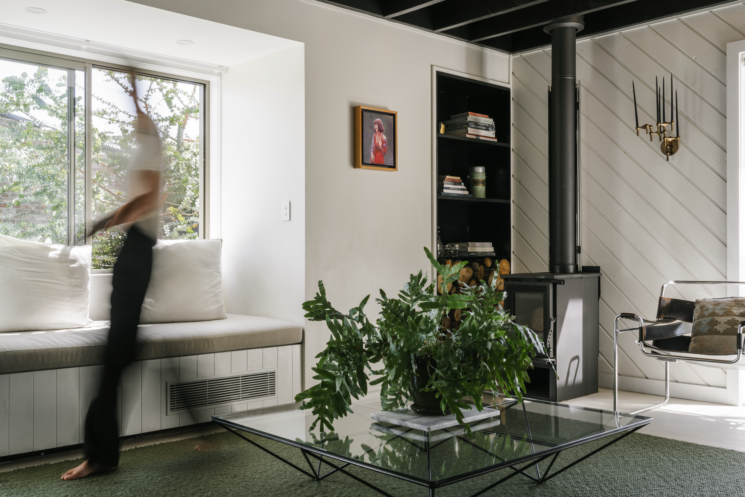 Living room with a person stretching by the window, white built-in window seat, green plant on glass coffee table, black wood stove, bookshelf, and modern chair.