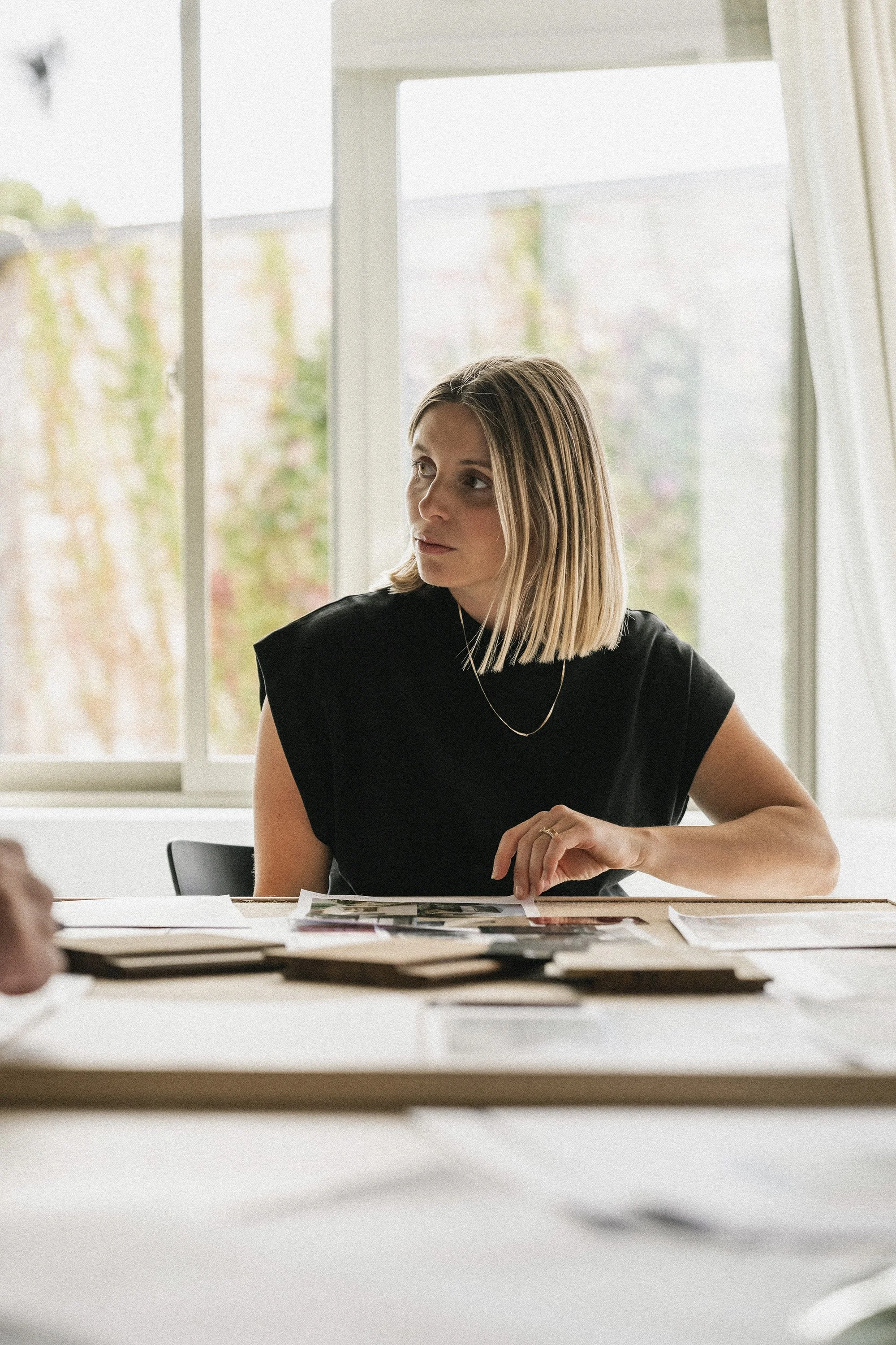 A woman with shoulder-length blonde hair in a black top sitting at a desk with papers, in front of a large window with a blurred outdoor background.