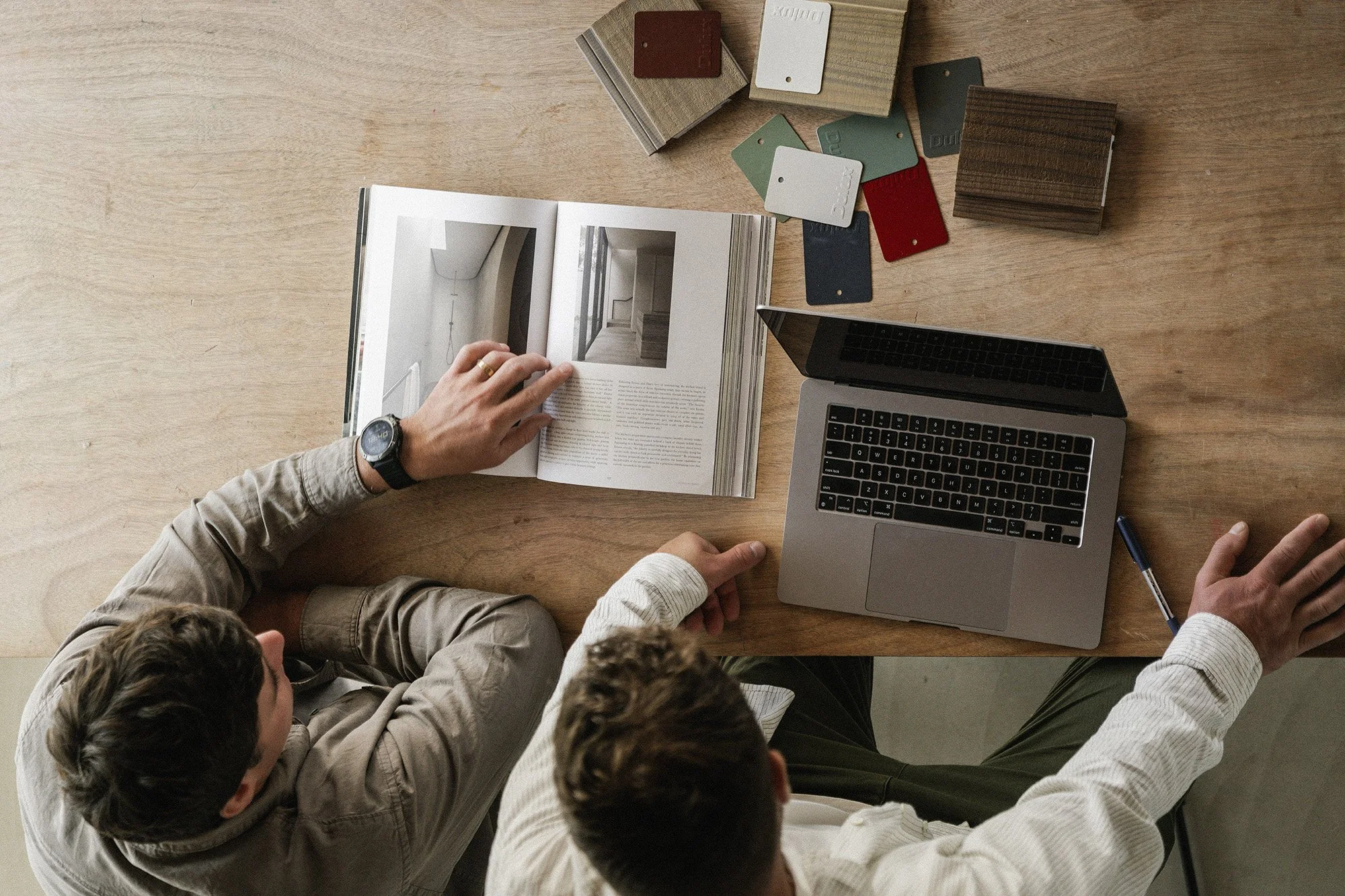 Top-down view of Tim and Hugh from Gather Group sitting at a wooden table, looking at an open magazine with interior design photos, with a laptop, pen, and color samples on the table.