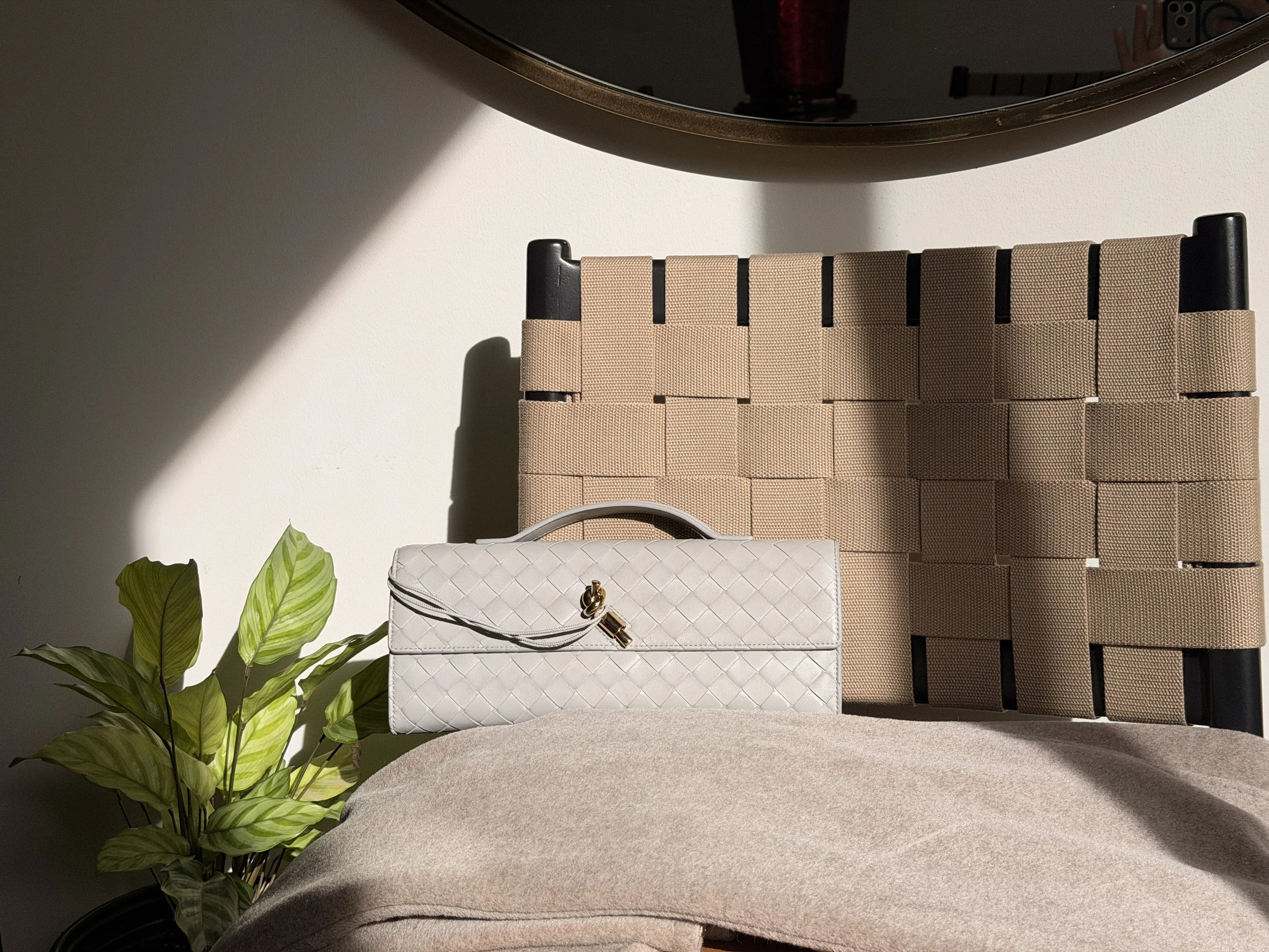 A beige handbag with a gold clasp on a beige upholstered surface, next to a green leafy plant, and a background consisting of a woven basket or wall decor, a wall mirror, and part of a person holding a camera.