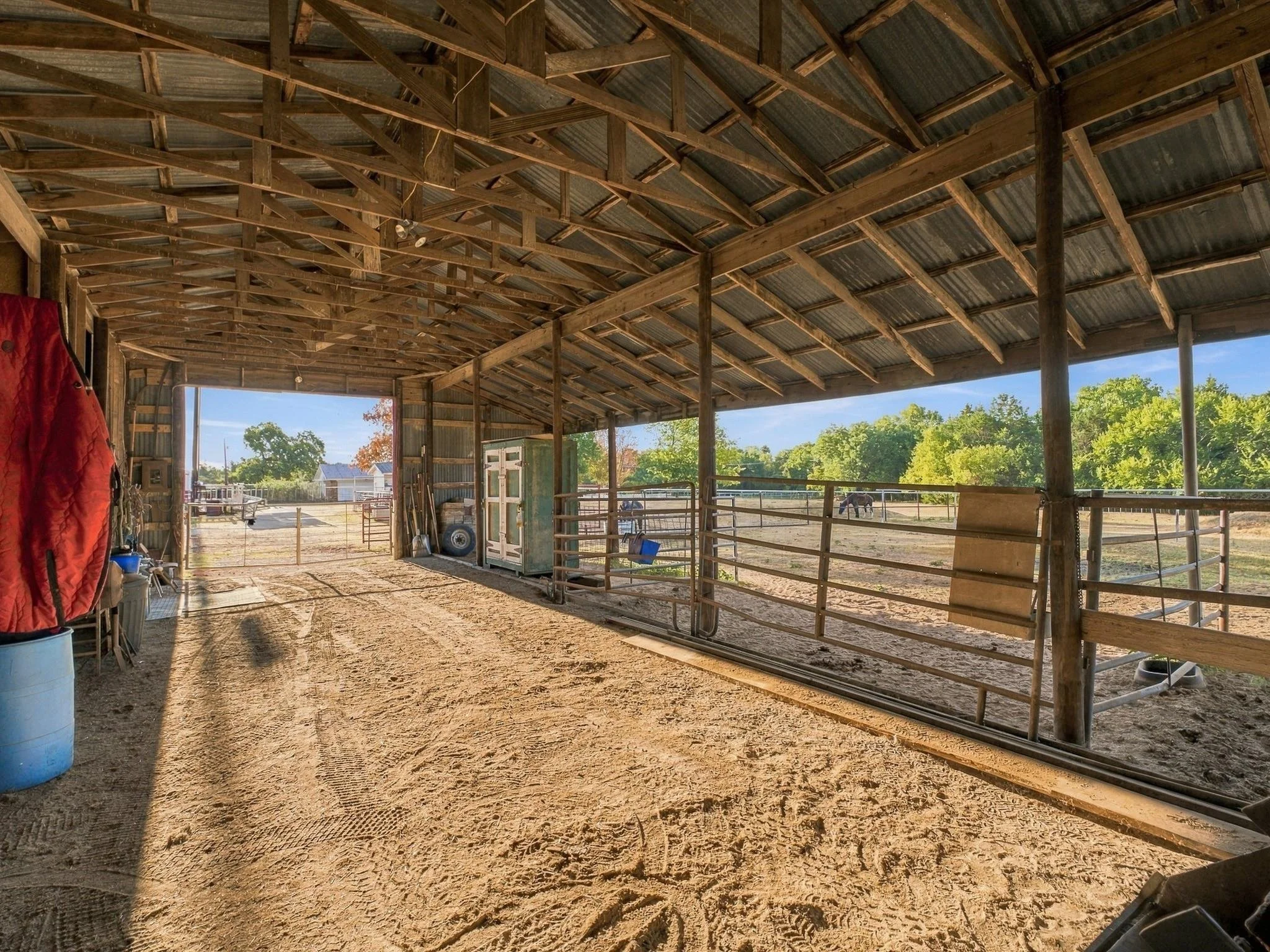 View inside a wooden barn with a dirt floor, overlooking an outdoor area with trees and blue sky. The barn has a high, angled roof with exposed wooden beams and metal roofing sheets.