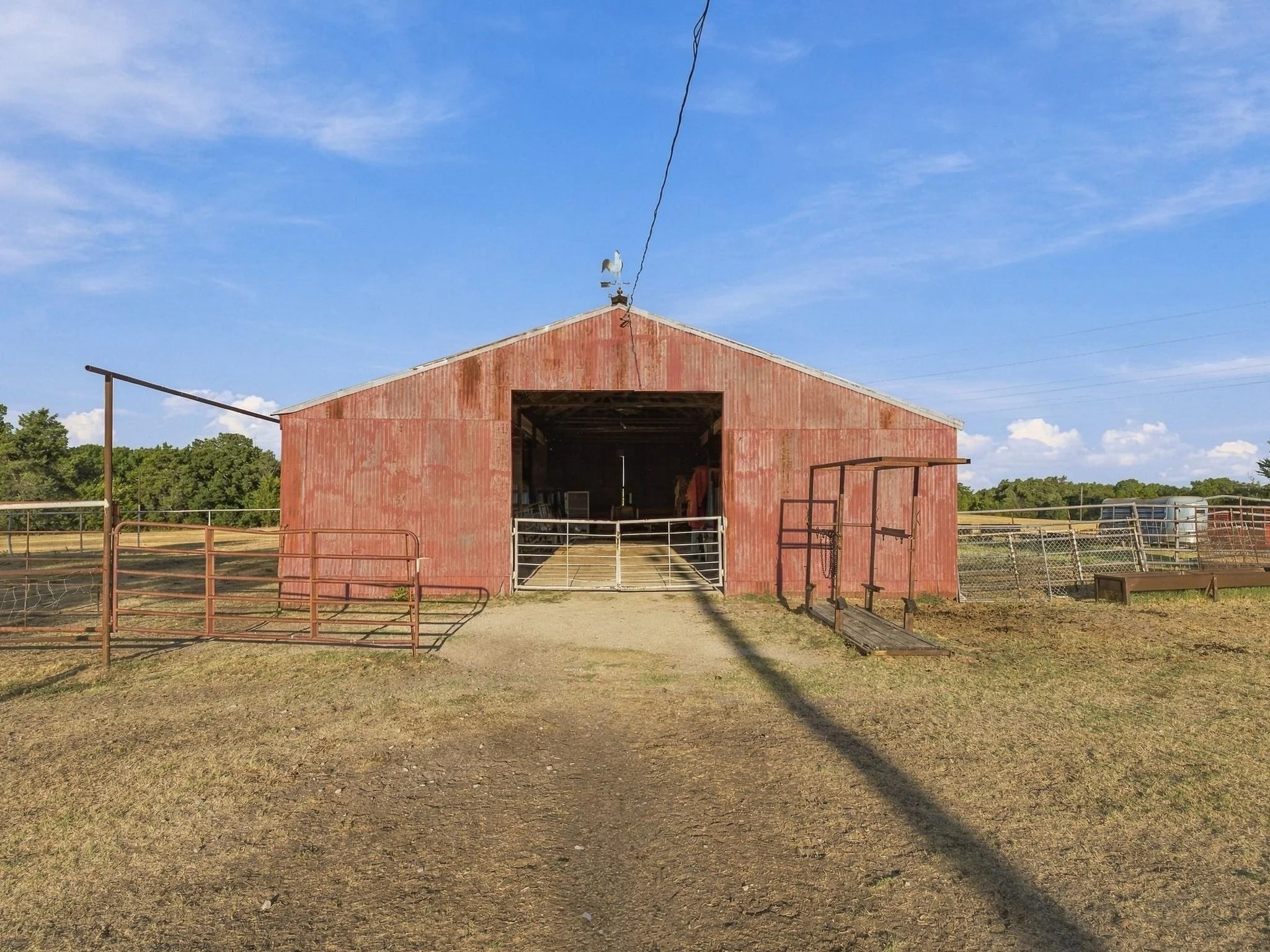 A red barn with a weathervane on top, surrounded by a fenced outdoor area under a clear blue sky with some wispy clouds.