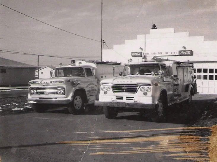 Two vintage fire trucks parked outside a fire department building with a sign that reads 'White River Twp Fire Dept' and Coca-Cola advertisements.
