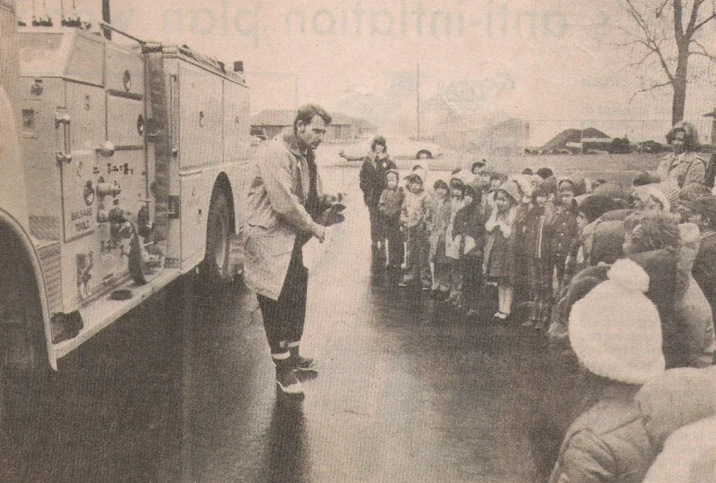 A firefighter standing next to a fire truck, talking to a group of children outdoors on a street, with some adults nearby.