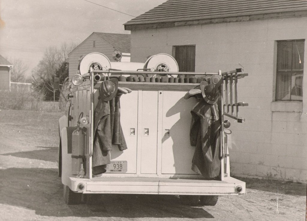 Back of vintage fire truck with two helmets, jackets, and firefighting equipment hanging, parked outside a building.