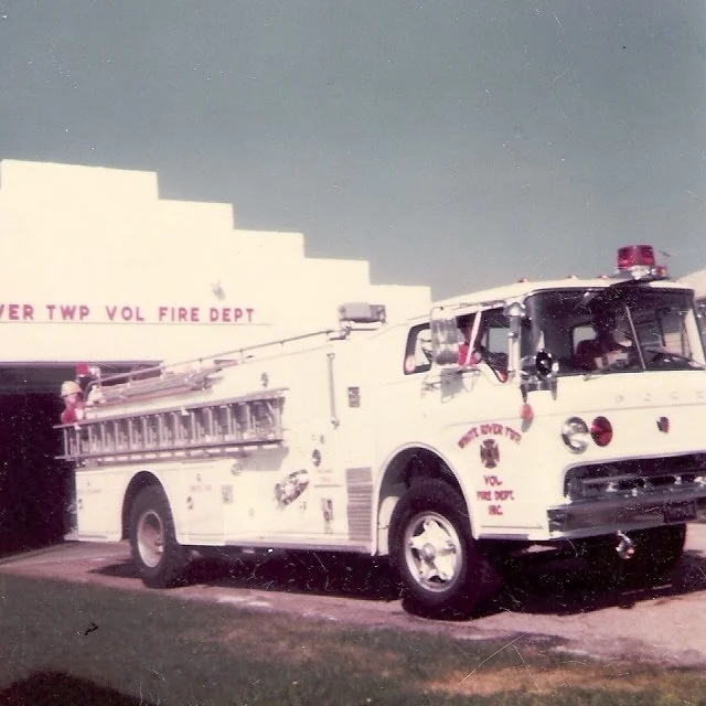 A vintage fire truck parked in front of a building with a sign that reads 'Over TWP Vol Fire Dept.' The fire truck is white with emergency lights on top and a ladder on the side.