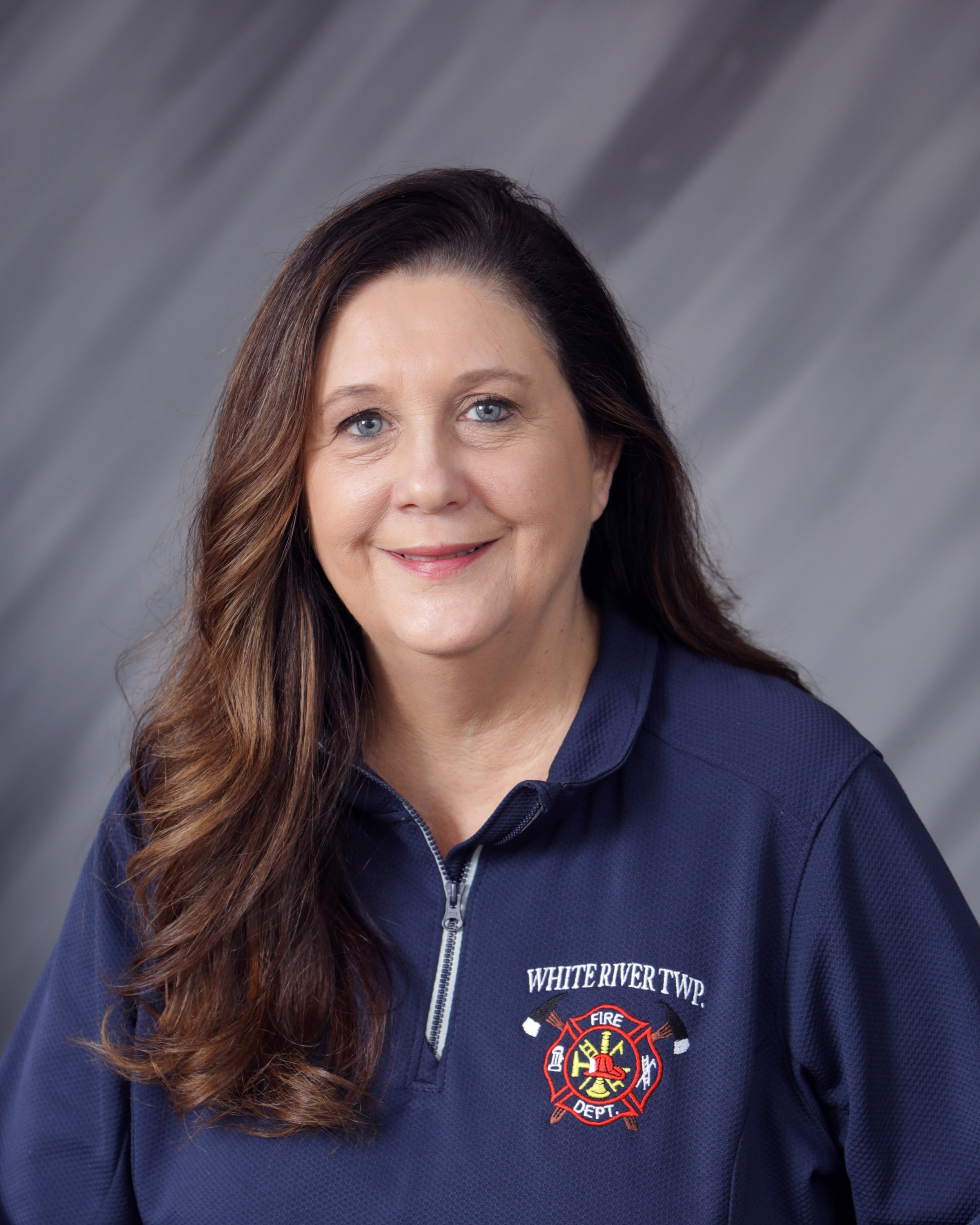 A woman with long brown hair and blue eyes smiling, wearing a navy blue firefighter uniform with an emblem and text 'WHITE RIVER TWP. FIRE DEPT.' on the chest, against a gray background.