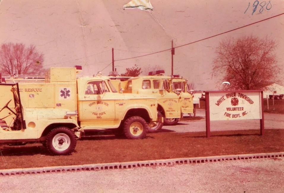 Three yellow fire rescue trucks parked in front of a sign that reads 'White River Township Volunteer Fire Department, Inc.' on a grassy area with trees in the background.