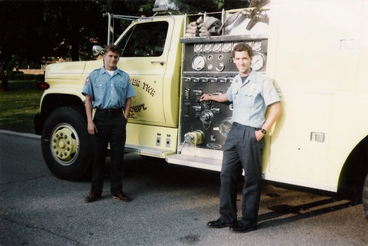Two firefighters in uniform standing next to a vintage yellow fire truck with a large control panel on the side.
