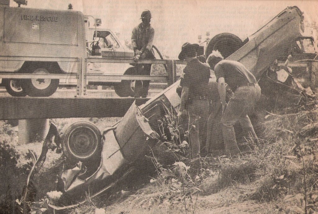 A car crash scene where a vehicle has flipped over on its side off the road with damage, and several people, including emergency responders, are inspecting the wreckage. A fire rescue vehicle is present nearby.