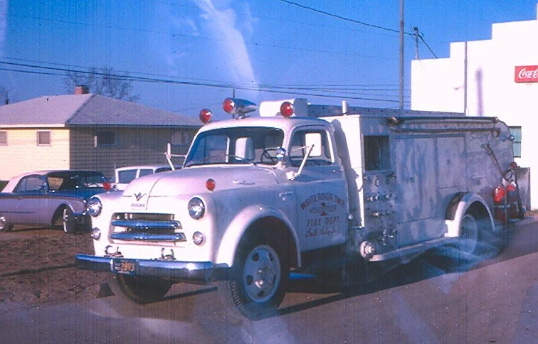 An old fire truck parked on a street with other cars and buildings in the background.